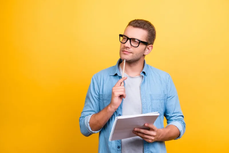 portrait with copy space of thoughtful, serious, bearded man in spectacles holding copybook and touching chin with pen with thoughtful expression over grey background
