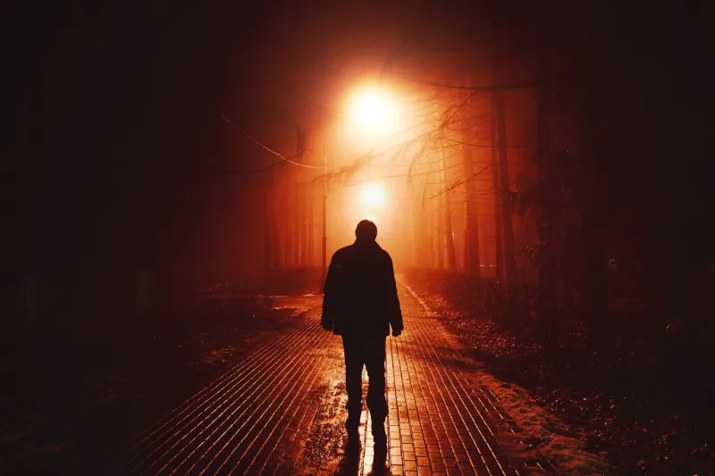 sad man alone walking along the alley in night foggy park back view