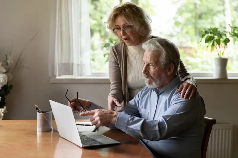 senior couple using app at home, studying new online app on modern tech device together mature man pointing at laptop screen, reading unexpected news, showing content to surprised middle aged wife