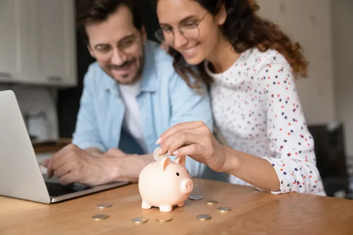 happy millennial family couple putting coins in piggybank, planning vacation or investments together, saving money for life insurance, managing future expenditures together using computer apps
