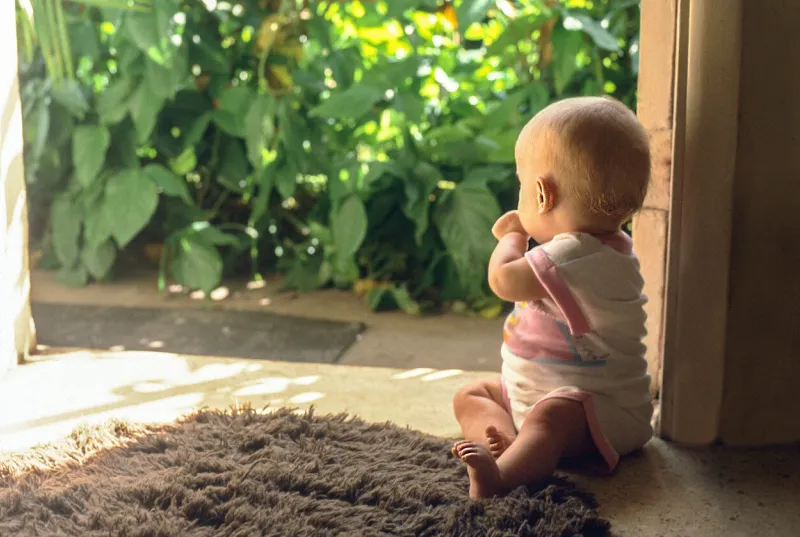 a horizontal image of an infant facing away from the camera looking out the open door there is greenery outside the infant is sucking her thumb and the sun is shining this image could evoke future, time, wonderment