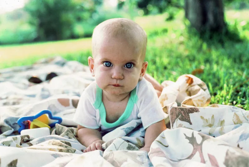 a young infant, old enough to hold her head up, lies on a blanket outside on the lawn in the shade the bright-eyed female child is looking directly at the camera with curiosity