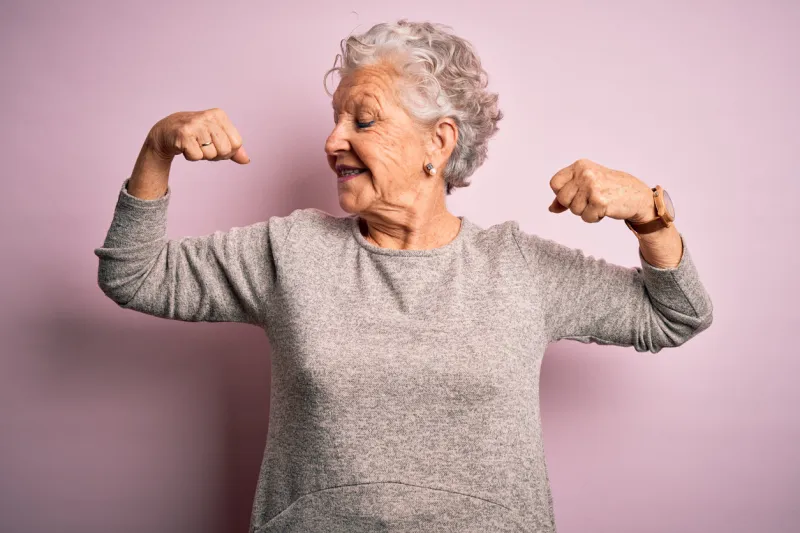senior beautiful woman wearing casual t-shirt standing over isolated pink background showing arms muscles smiling proud fitness concept