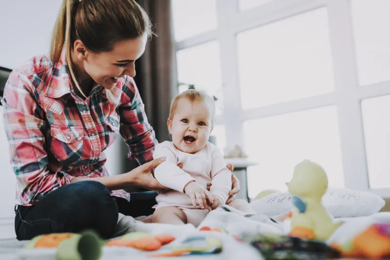 mother sits with child on floor and holding doll young smiling mother wears casual clothes playing toys with cute caucasian baby on floor near large panoramic window in living room