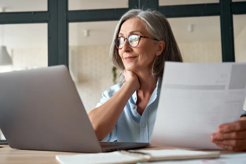 smiling mature middle aged business woman using laptop working on computer sitting at desk happy old businesswoman hr holding cv interviewing distance applicant, senior seeker searching job online