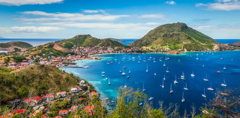 colorful image of guadeloupe terre de haut bay and town with buildings along the coastline small ships anchored in port cruise destination green mountains in the background blue sky and some white clouds