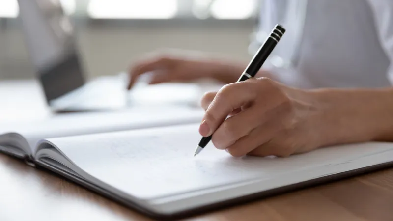 female professional doctor hand making notes in medical journal using laptop computer sitting at desk woman physician, nurse or pharmacist wearing white coat writing in paper notebook close up view