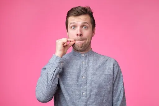 young man showing a sign of closing mouth and silence gesture doing like closing his mouth with a zipper on isolated pink background