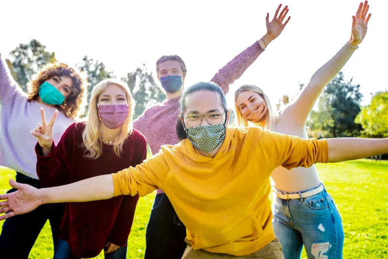 happy group of multiracial people covered by face masks having fun outside - new normal friendship concept with young friends with hands up outdoor