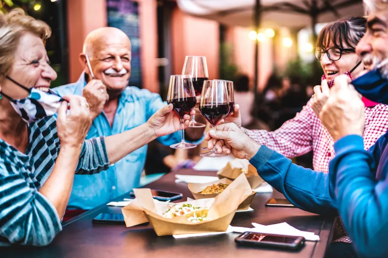 senior people toasting wine at restaurant bar wearing open face masks - new normal life style concept with happy mature friends having fun together at garden party - vivid filter with focus on glasses