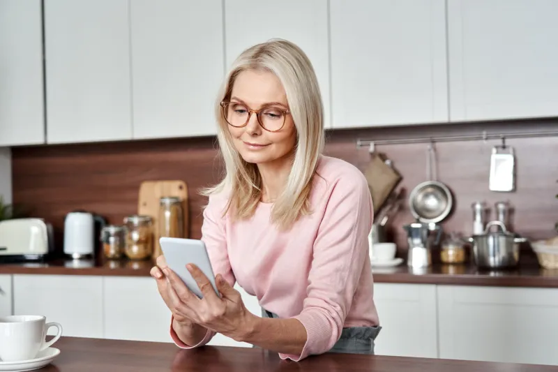 middle aged 50 years old woman using apps ordering buying food on smartphone sitting in kitchen at home mature older lady holding mobile phone texting messages, browsing online services