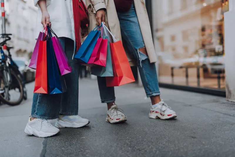 look what we bought close up of two girl in jeans and sneakers holding colorful shopping bags