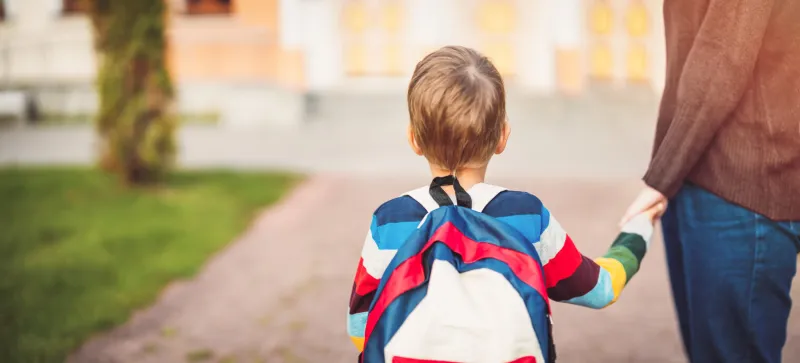 child with rucksack and with mother in front of a school building concept of motherhood and beginning of primary school