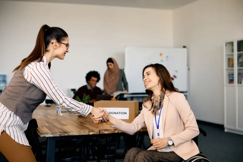 happy charity owner in wheelchair handshaking with a businesswoman during a meeting at charitable foundation