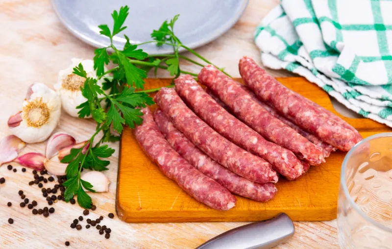 raw spicy longaniza sausages from minced pork prepared for frying lying on wooden board with knife, garlic, peppercorns and fresh parsley traditional spanish specialty