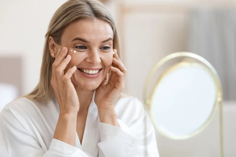happy beautiful blonde middle aged lady in silky bathrobe using moisturizing eye cream, looking at mirror and smiling, enjoying face care routine, closeup shot dark circles, eye wrinkles treatment