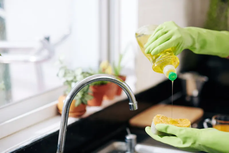 closeup image of woman in latex gloves pouring dish soap on sponge and washing dishes