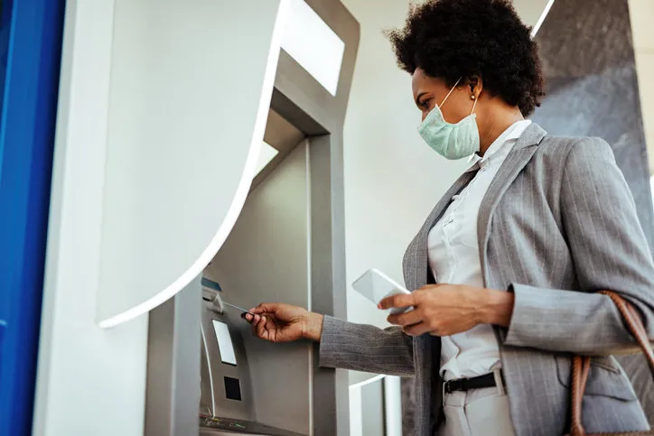 low angle view of african american businesswoman inserting credit card and withdrawing cash at atm while wearing protective mask on her face
