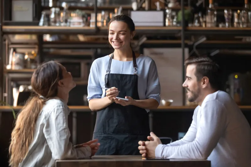 smiling waitress wear apron hold notepad pen take order talk to clients serving restaurant guests couple choosing food drinks menu sit at cafe coffeehouse table, waiting staff, good customer service