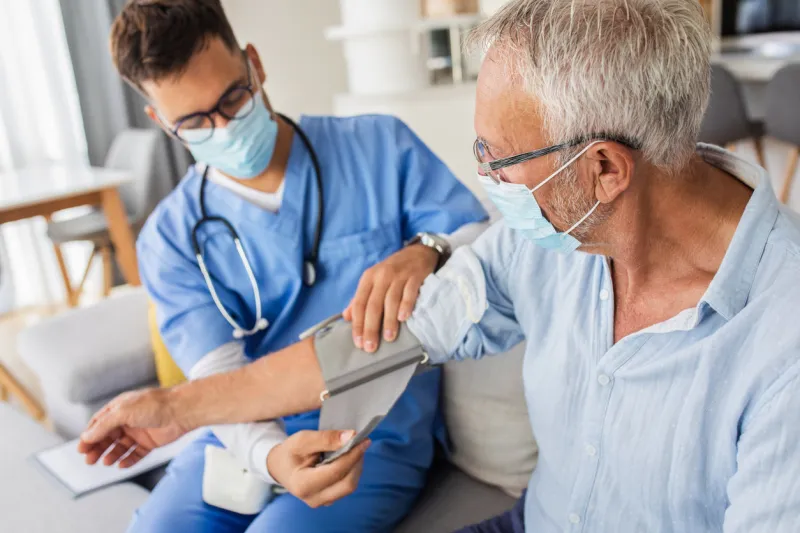 male nurse measures blood pressure to senior man with mask while being in a home visit