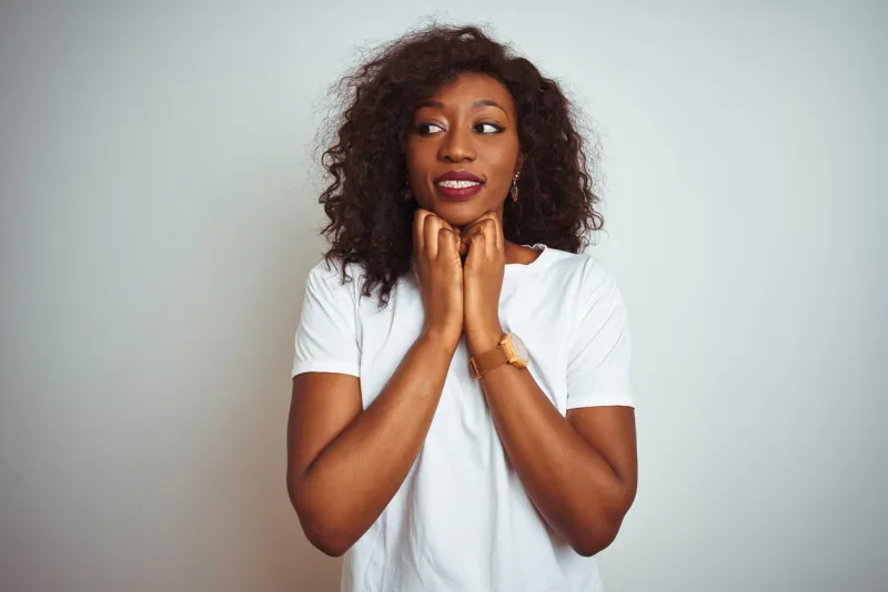 young african american woman wearing t-shirt standing over isolated white background laughing nervous and excited with hands on chin looking to the side