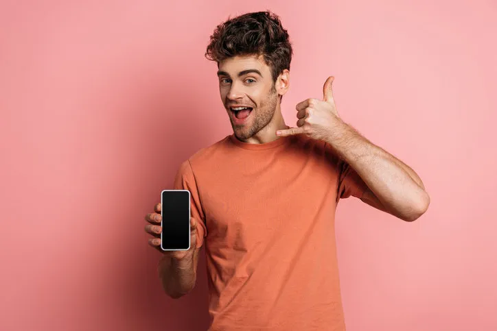 cheerful young man showing lets drink gesture while holding smartphone with blank screen on pink background