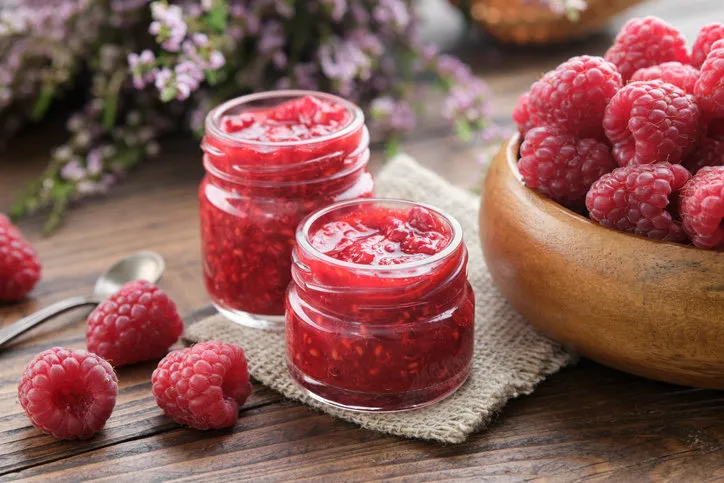two small jars of raspberries jam, wooden bowl of fresh red raspberries on kitchen table
