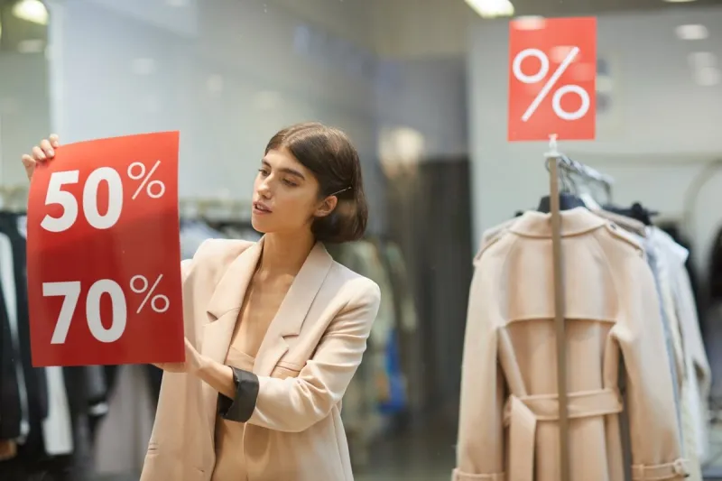 waist up portrait of beautiful woman hanging red sale signs on window display in clothes store, copy space