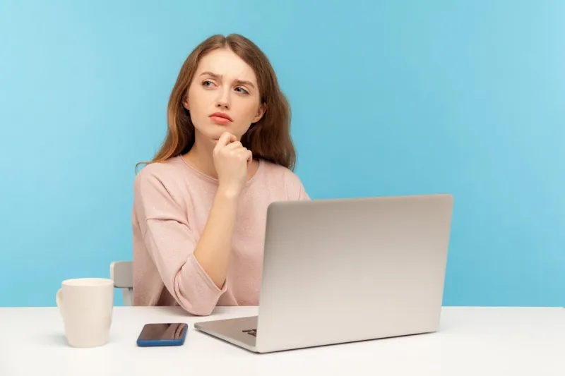 need to think over business plan pensive woman in casual clothes sitting with thoughtful confused expression while working on laptop at home office indoor studio shot isolated on blue background