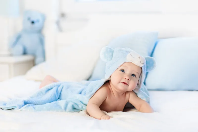 baby boy wearing diaper and blue towel in white sunny bedroom newborn child relaxing in bed after bath or shower nursery for children textile and bedding for kids new born kid with toy bear