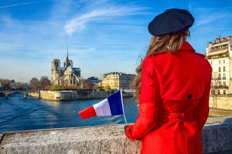 bright in paris seen from behind modern traveller woman in red trench coat on embankment near notre dame de paris in paris, france with french flag