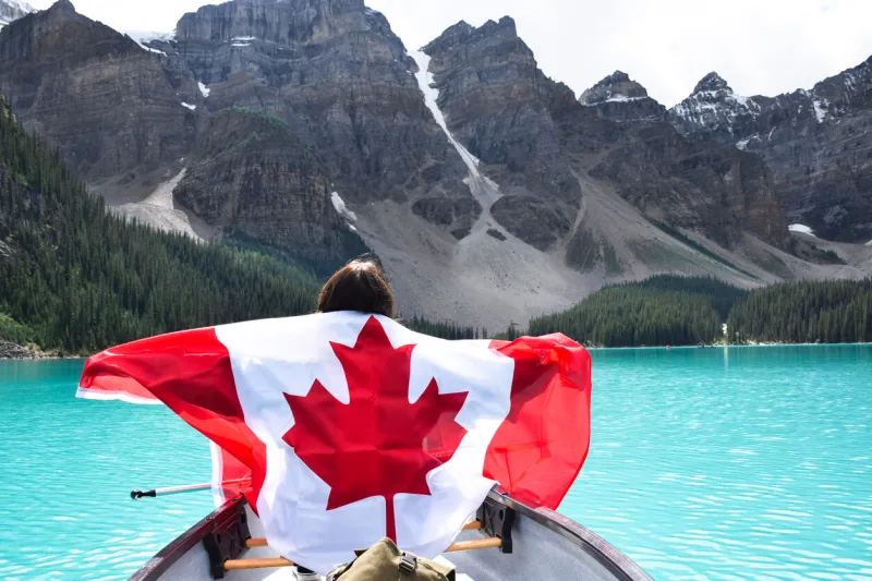young girl in a canoe from behind holding canadian flag spreading her arms, surrounded by turqouise blue lake moraine and mountains