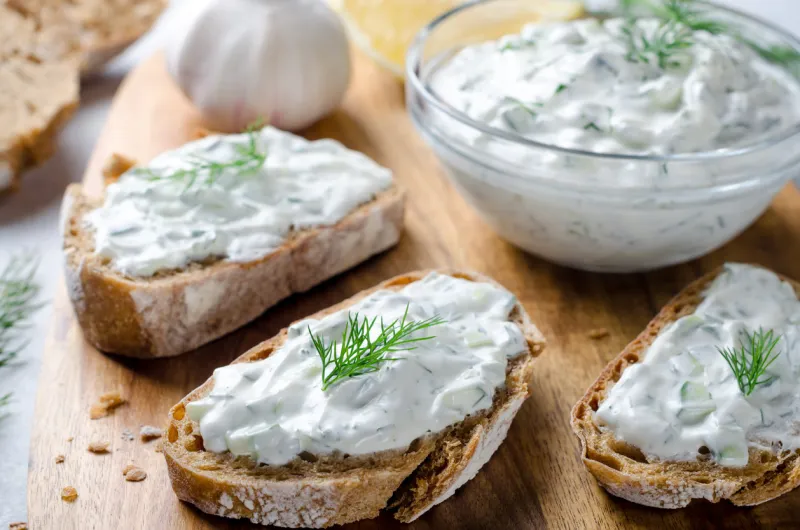 homemade greek tzatziki sauce in a glass bowl with sliced bread on a wooden board close-up, horizontal image, selective focus on bread