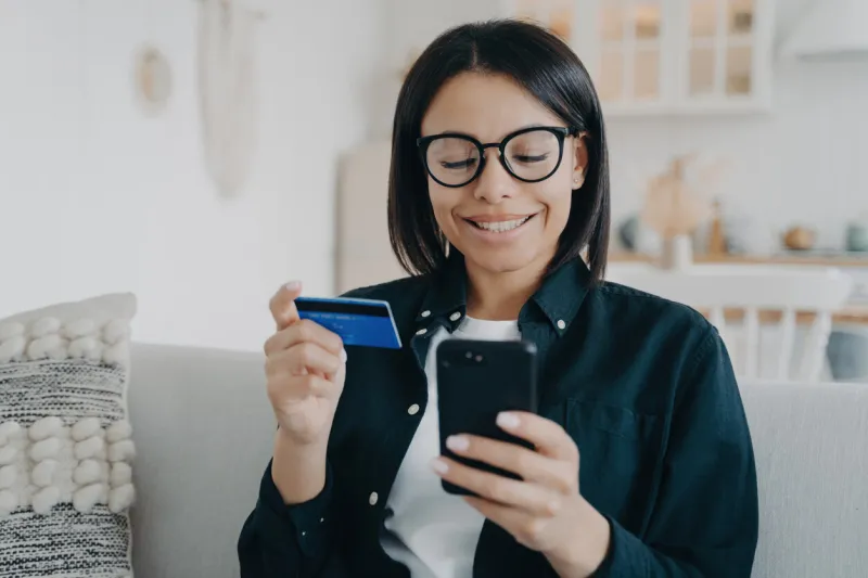 smiling female holding bank credit card and smartphone young woman pays for purchases in online store using banking service, shopping, making secure payment, sitting on sofa at home e-commerce
