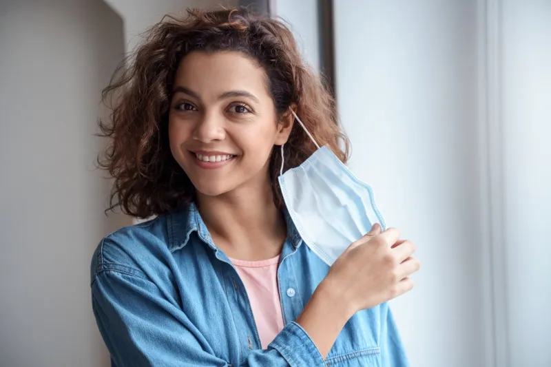 happy hispanic young woman takes off protective mask and looking at camera indoors while corona virus pandemic quarantine, and social distance concept