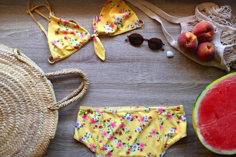 yellow floral swimming suit, sunglasses, mesh tote with fresh peaches, seashell, watermelon half and wicker bag on wooden background summer essentials, flat lay