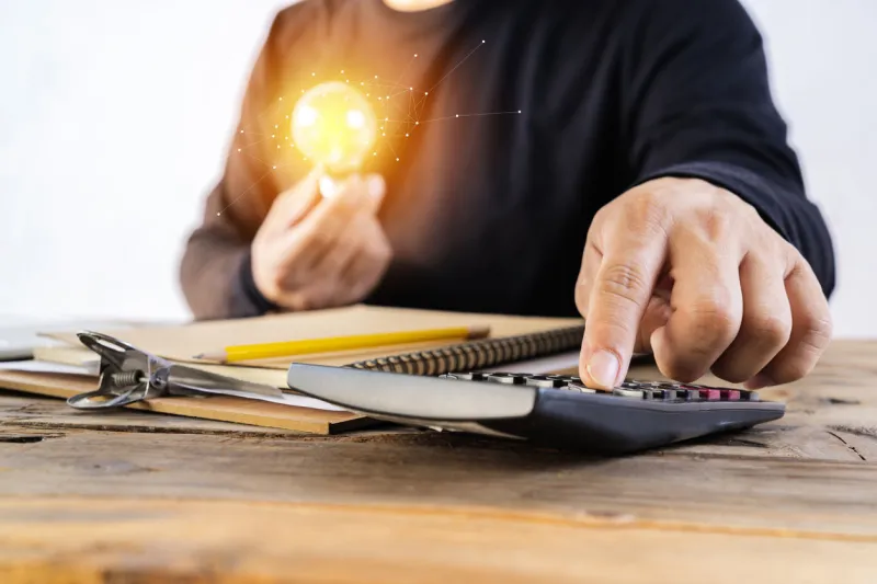 human hand holds a light bulb and presses a calculator to calculate the company energy use numbers saving energy and accounting finance concept
