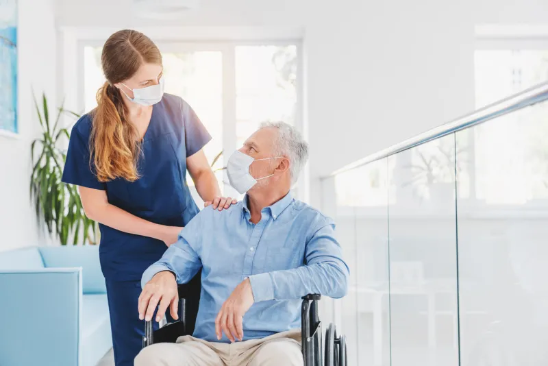 young woman nurse explaining information to man patient in wheelchair in medical face mask while talking together in hospital epidemic and virus concept