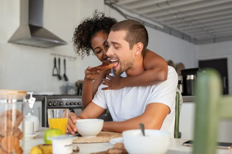 african american woman embracing from behind her boyfriend and feeding him with brown bread and jam multiethnic couple enjoying breakfast at home beautiful laughing woman feeding young man in kitchen and having fun together in the morning