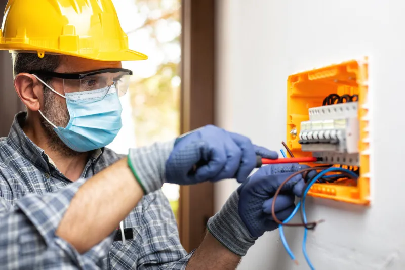 electrician at work on an electrical panel protected by helmet, safety goggles and gloves, wear the surgical mask to prevent the spread of coronavirus construction industry covid-19 prevention