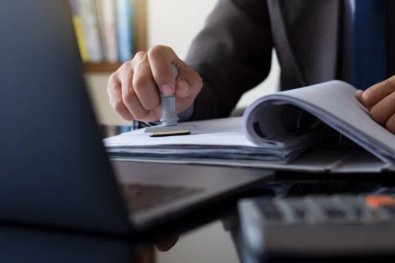businessman in suit hand stamping rubber stamp on document in file folder with laptop computer on the desk at office authorized allowance permission approval concept