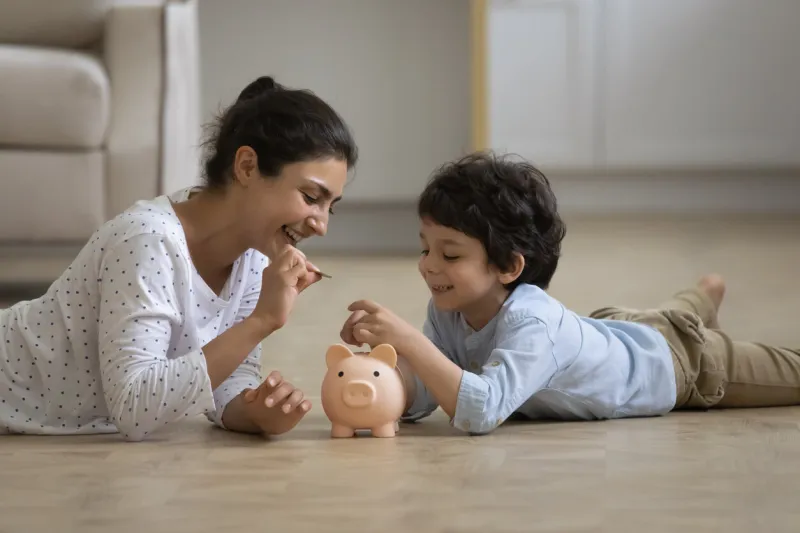 caring young asian indian mother teaching little kid son saving money or planning future purchases, putting coins in piggybank, lying on heated floor, financial education for children concept
