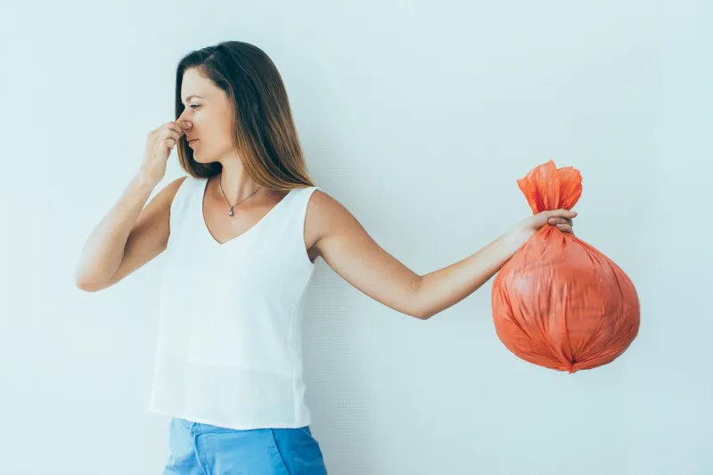 displeased young woman with pinched face holding garbage bag and holding her nose unpleasant smell concept