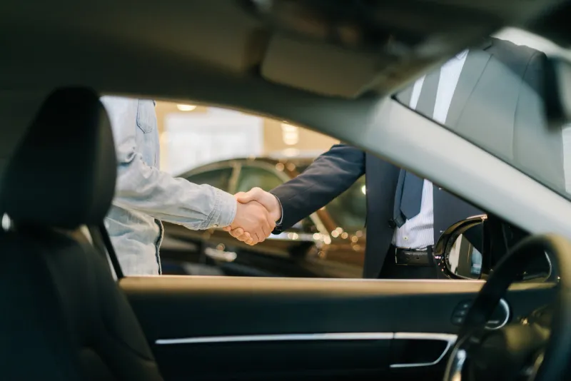 buyer of car shaking hands with seller in auto dealership, view from interior of car close-up of handshake of business people concept of choosing and buying new car at showroom