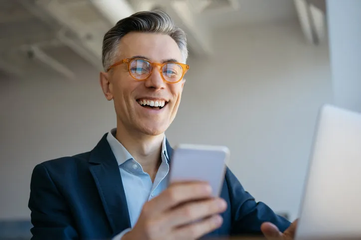 smiling mature man using laptop computer, holding mobile phone sitting in modern office, successful business portrait of handsome emotional freelancer receive payment, working from home