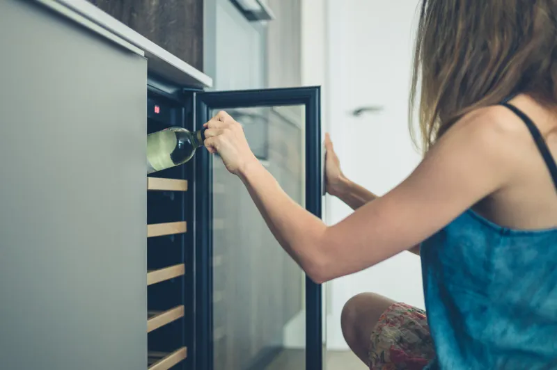 a young woman is getting a bottle of white wine from her wine cooler at home