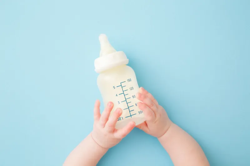 infant hands holding bottle of milk on light blue floor background feeding time pastel color closeup point of view shot top down view