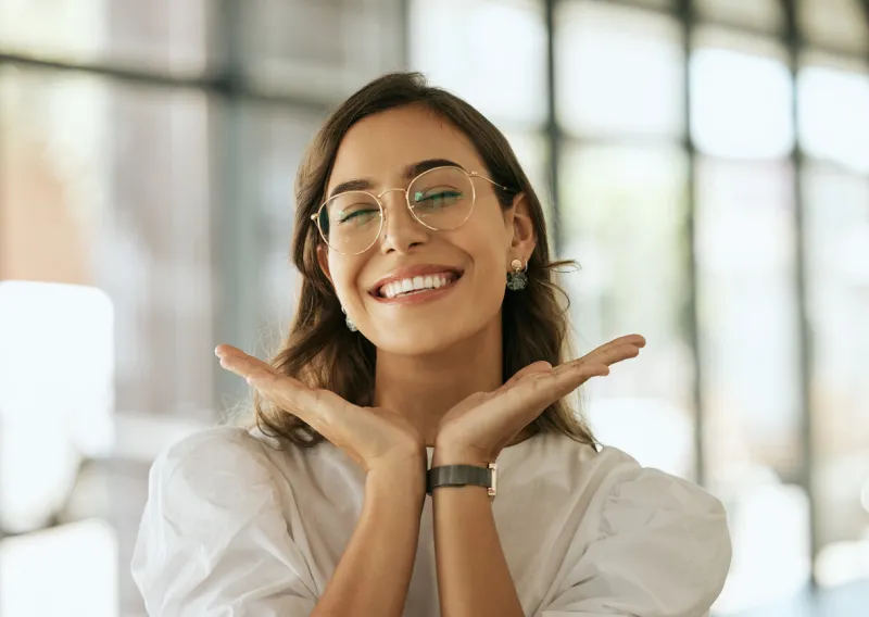 cheerful business woman with glasses posing with her hands under her face showing her smile in an office playful hispanic female entrepreneur looking happy and excited at workplace