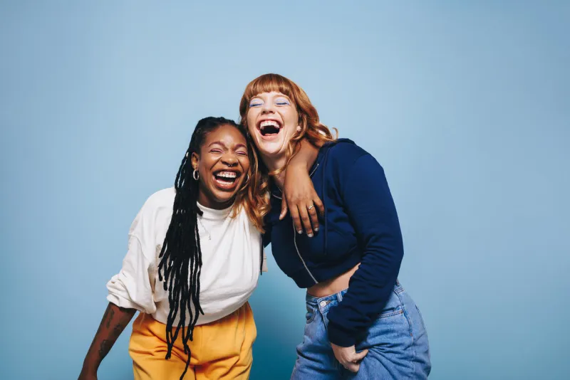 interracial best friends laughing and having a good time together in a studio happy young women enjoying themselves while standing against a blue background two vibrant female friends making memories