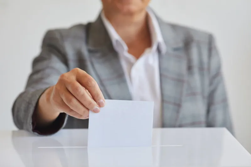 front view close up of female hand putting vote bulletin in ballot box against white background on election day, copy space